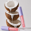 Drei halbierte Kokosnüsse, aufeinandergestapelt, aus denen Kokosmilch tropft; daneben liegen zwei Tuben „BLONDESISTER 3 IN 1 CUTE LIPPY LIP OIL“.