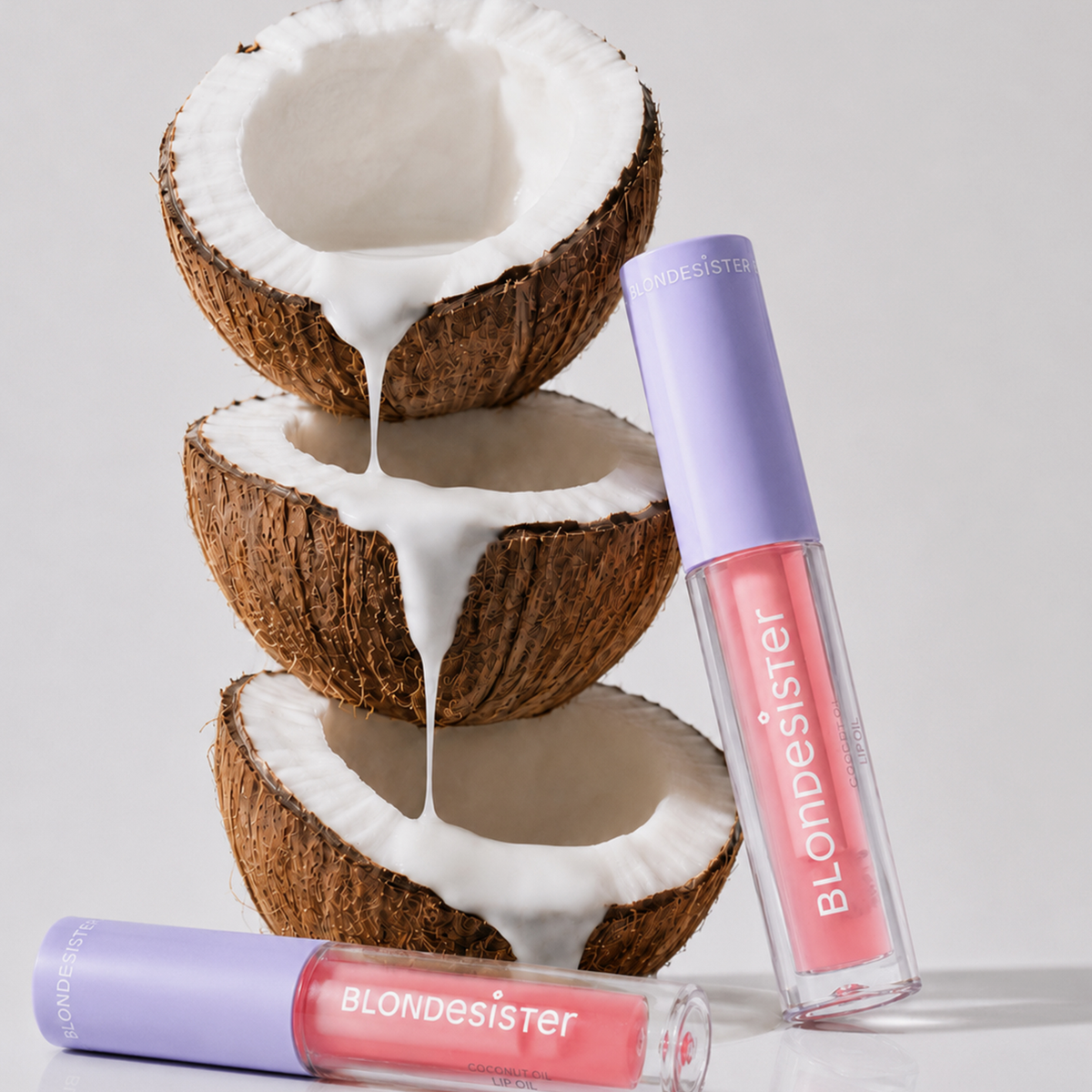 Drei halbierte Kokosnüsse, aufeinandergestapelt, aus denen Kokosmilch tropft; daneben liegen zwei Tuben „BLONDESISTER 3 IN 1 CUTE LIPPY LIP OIL“.