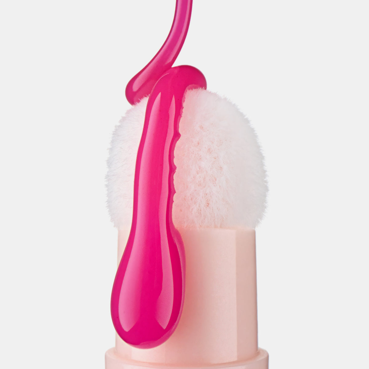 Nahaufnahme eines weißen Kissenschwamms, auf den leuchtend pinkes Lip Oil tropft, Produkt in elegantem Nude-Gehäuse vor weißem Hintergrund.