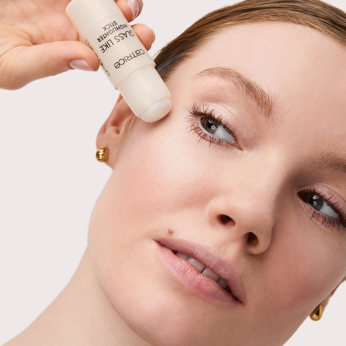 Eine Frau trägt den Catrice Cosmetics Glass Like Highlighter Stick 010 auf ihren Wangenknochen in der Nähe ihres Auges auf.