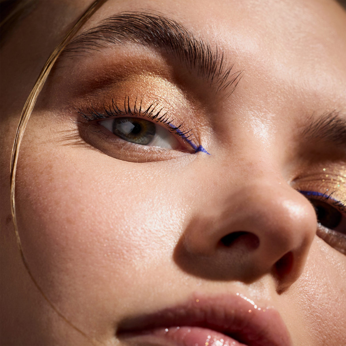 Nahaufnahme einer Frau, mit goldenen Farbtönen und blauem Eyeliner; strahlende Haut und glänzende Lippen.