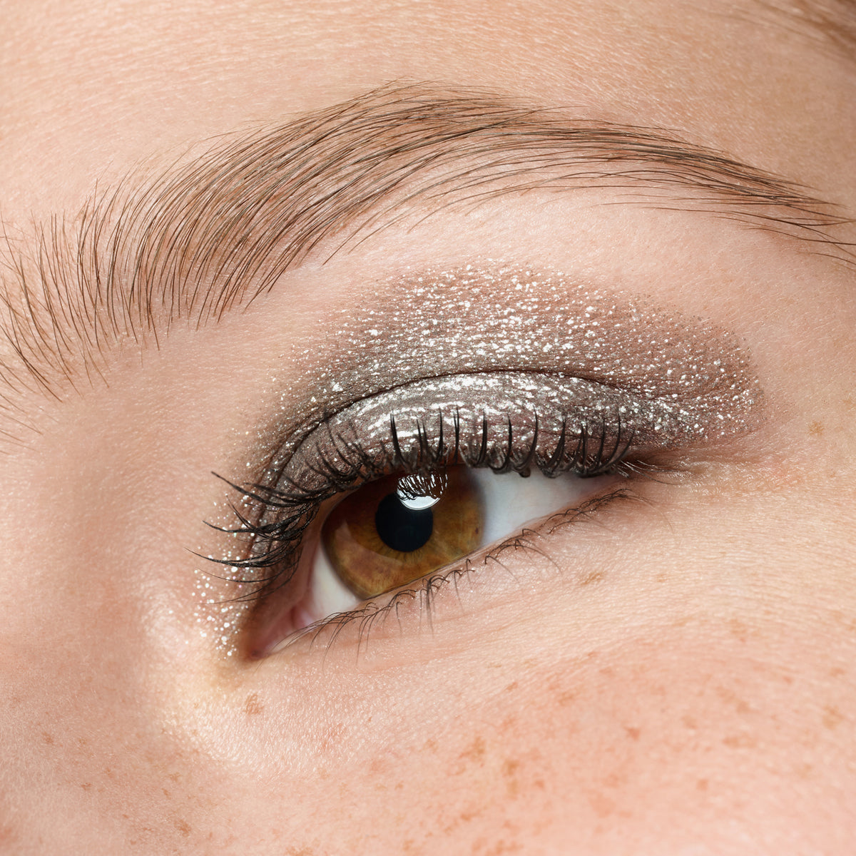 Eine Nahaufnahme eines Auges mit brauner Iris und langen Wimpern, mit sommersprossiger Haut und silbernem Glitzerlidschatten.