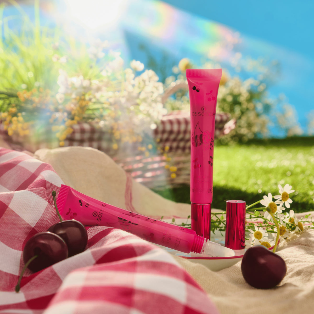Zwei SO CHERRY Happy Peptide Lipgloss-Tuben mit Kirschdesign liegen auf einer Picknickdecke, umgeben von Blumen im hellen Sonnenlicht.