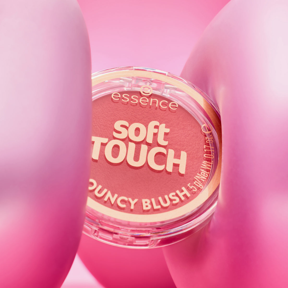 Ein runder Essence soft TOUCH BOUNCY BLUSH Behälter wird zwischen zwei rosafarbenen, ballonartigen Formen gehalten.