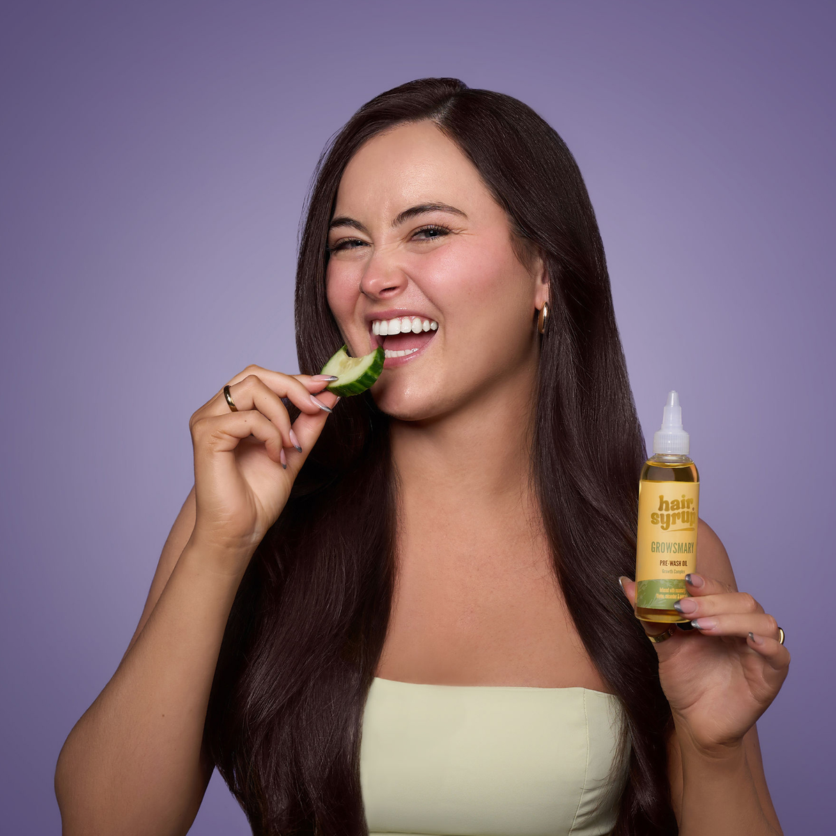 Lächelnde Frau mit langen dunklen Haaren hält eine Gurkenscheibe und eine Flasche Growsmary by Hair Syrup vor einem lila Hintergrund.