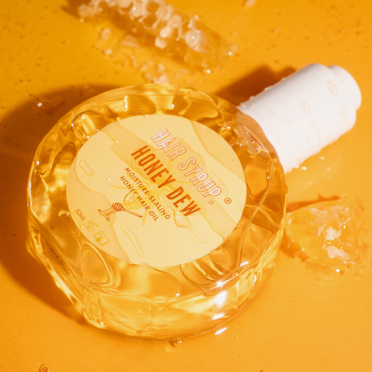 Eine runde Flasche mit Hair Syrup Honeydew Oil steht auf einer honigfarbenen Fläche mit Wabenstücken in der Nähe.