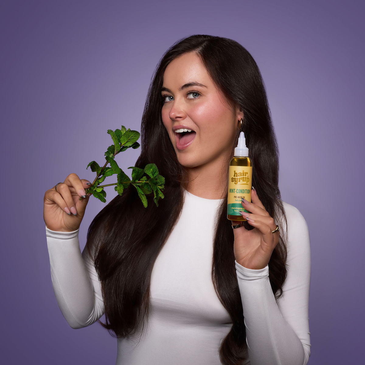 Eine Frau mit langen dunklen Haaren hält einen Minzzweig und eine Flasche Hair Syrup's Mint Condition vor einem lila Hintergrund.