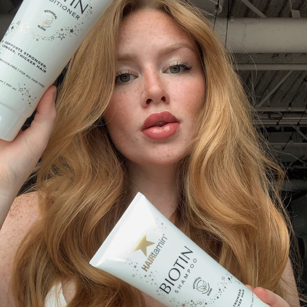 Frau mit langen, welligen blonden Haaren hält Hairtamin Biotin Shampoo und Conditioner in den Händen, Fokus auf glänzendes, gepflegtes Haar.