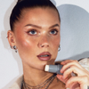 Frau mit strahlender Haut und mehreren Ohrringen trägt LH Cosmetics Artistick Highlighter auf ihre Lippen auf.