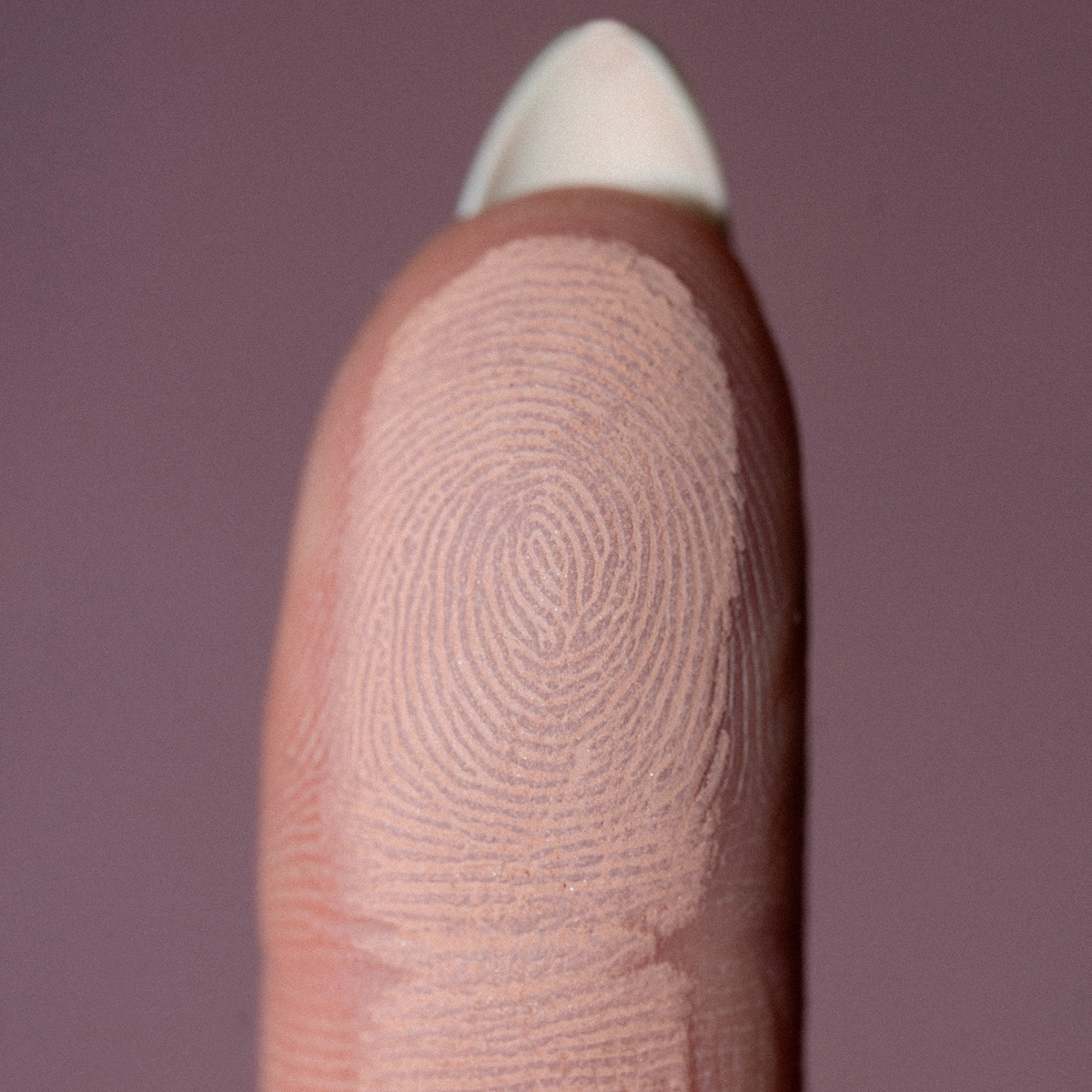 Eine Nahaufnahme einer Fingerkuppe mit detaillierten Fingerabdruckrillen, die The Single von LH Cosmetics auf einem unscharfen lila Hintergrund zeigt.