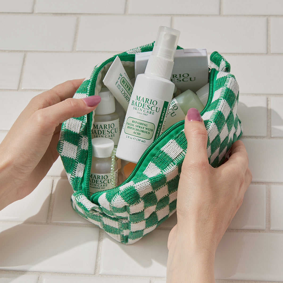 Hände halten eine grün karierte Kosmetiktasche mit Produkten von Mario Badescu auf einem weiß gekachelten Hintergrund