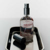 Eine Flasche MERME Antioxidant Mist With Rose Quartz mit abgenommenem Verschluss steht auf einem Metalltablett vor einem hellen Hintergrund.