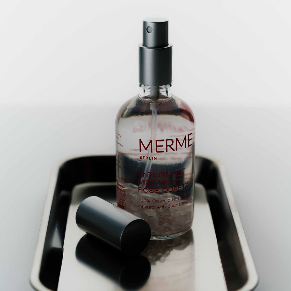 Eine Flasche MERME Antioxidant Mist With Rose Quartz mit abgenommenem Verschluss steht auf einem Metalltablett vor einem hellen Hintergrund.