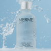 Glasflasche des MERME Oxygen Water 4-Wochen-Sets mit silbernem Deckel mit plätscherndem Wasser vor hellblauem Hintergrund.