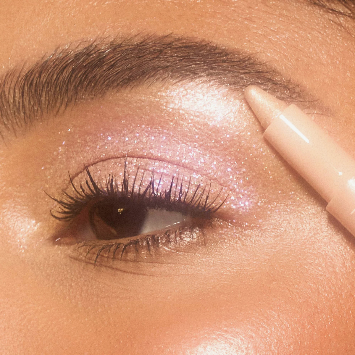 Nahaufnahme eines Auges mit rosa-lila Glitzer-Look. Ein heller Lidschattenstift setzt strahlende Highlights am Brauenbogen für einen leuchtenden Effekt.