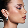 Profilaufnahme einer Person mit grafischem Augen-Make-up in Lila-, Rosa- und Brauntönen sowie markanten metallischen Ohrringen.