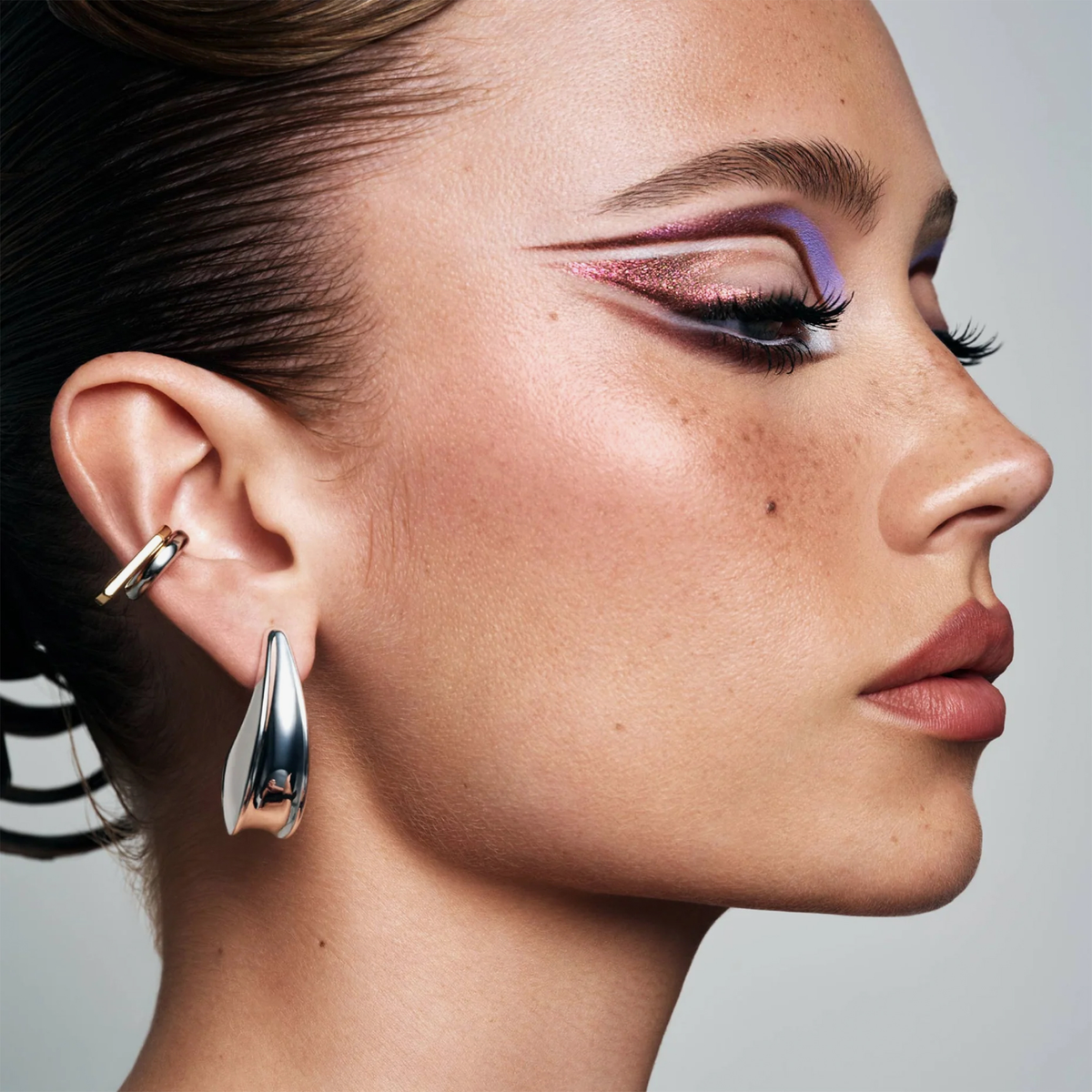Profilaufnahme einer Person mit grafischem Augen-Make-up in Lila-, Rosa- und Brauntönen sowie markanten metallischen Ohrringen.