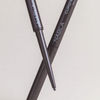 Zwei NABLA Cosmetics Cupids Arrow Micro Eyeliners in Dunkelbraun, gekreuzt, einer mit freiliegender Spitze, auf hellem Grund.