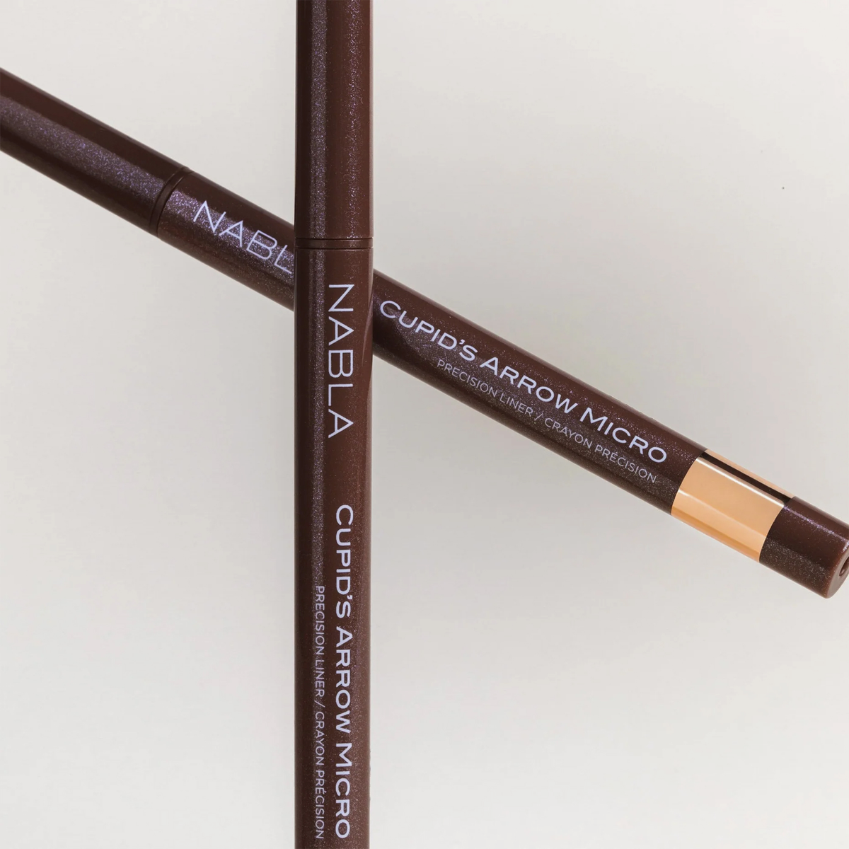 Zwei braune NABLA Cosmetics Cupids Arrow Micro Eyeliner Stifte gekreuzt auf einem hellen Hintergrund.