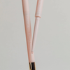 Zwei rosafarbene NABLA Cosmetics Cupids Arrow Micro Augenbrauenstifte, einer ohne Kappe, um die Präzisionsspitze zu zeigen, auf einem hellen Hintergrund.