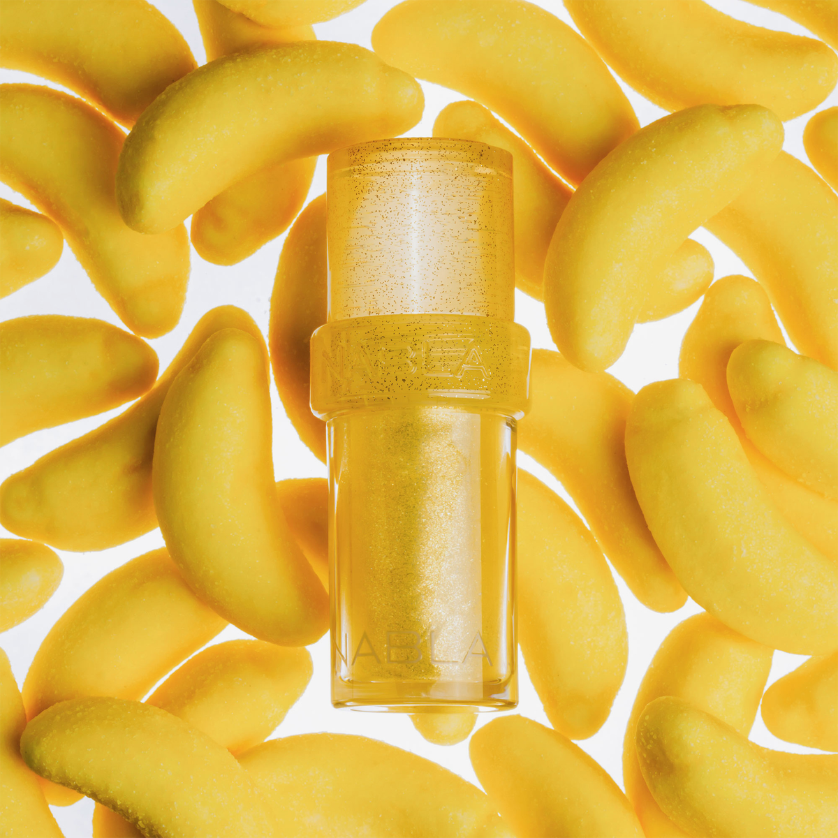 Zwischen kleinen, leuchtend gelben, bananenförmigen Gegenständen steht eine Tube „Lip Candy Oil“ von NABLA Cosmetics.