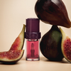 Das „Lip Candy Oil“ von NABLA Cosmetics steht zwischen halbierten und ganzen Feigen vor einem beigen Hintergrund.