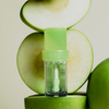 Das „Lip Candy Oil“ von NABLA Cosmetics steht vor einem Stapel grüner Äpfel, von denen einige ganz und andere in Scheiben geschnitten sind.