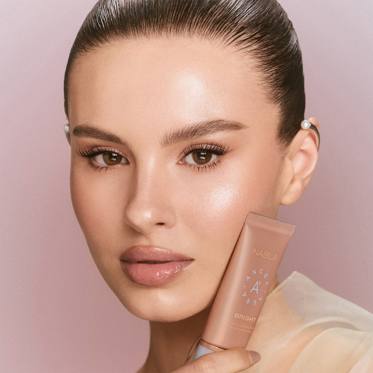 Eine Frau mit strahlender Haut hält eine Tube Angel Aura Bright des NABLA Cosmetics Sun-Kissed Glow Set vor einem zartrosa Hintergrund.