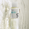 Eine klare Flasche des numbuzin No.5+ Vitamin Boosting Essence Toner steht vor einem wässrigen, strukturierten Hintergrund.