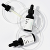 Zwei schwarze Tropfflaschen mit numbuzin No.5+ Vitamin Concentrated Serum sind in Petrischalen auf weißem Hintergrund ausgestellt.
