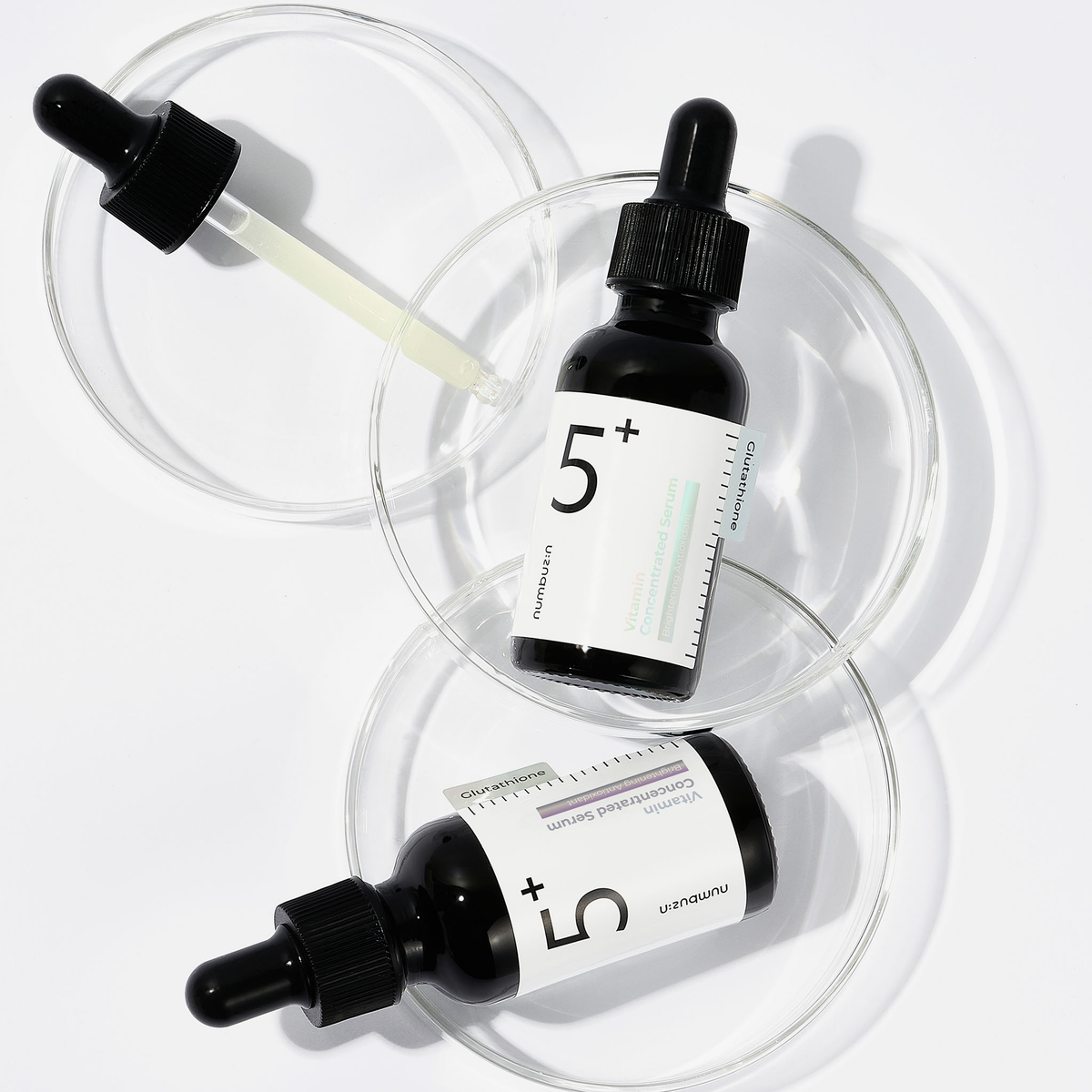 Zwei schwarze Tropfflaschen mit numbuzin No.5+ Vitamin Concentrated Serum sind in Petrischalen auf weißem Hintergrund ausgestellt.