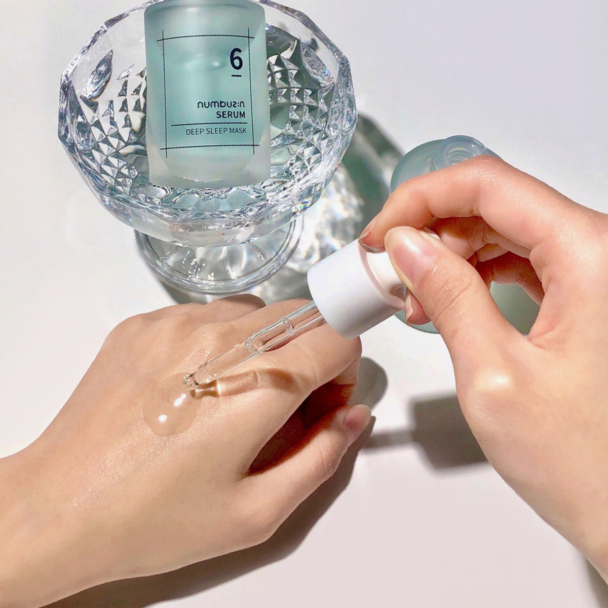 Eine Hand gibt mit einer Pipette das „Numbuzin No. 6 Deep Sleep Mask Serum“ auf die andere Hand; die Serumflasche steht daneben in einer Glasschale.