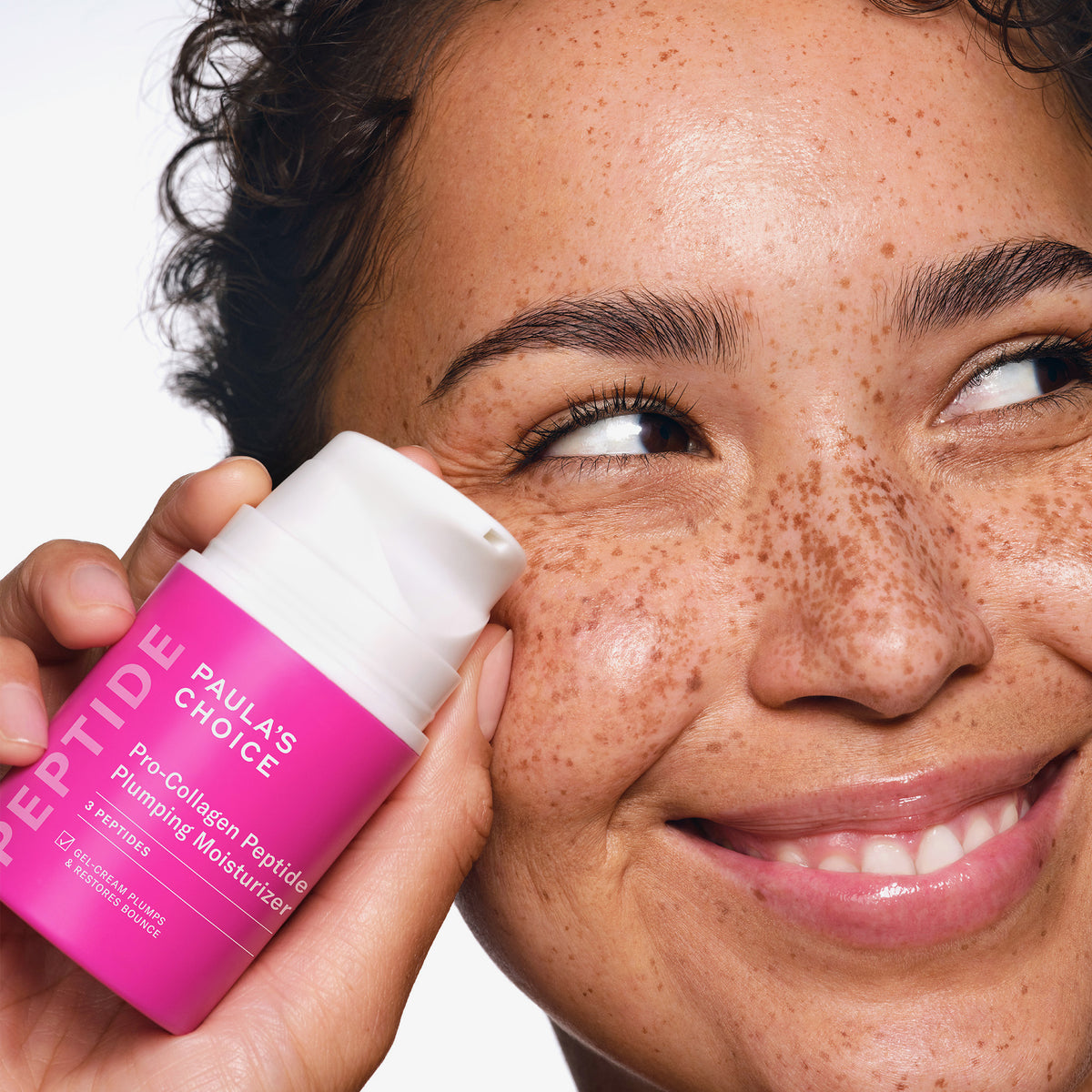 Eine lächelnde Person mit Sommersprossen hält eine Flasche Paula's Choice Pro-Collagen Peptide Plumping Feuchtigkeitscreme an ihre Wange.