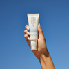 Eine gepflegte Hand hält das „Philip Kingsley Finishing Touch Frizz Fighting Gloss“ vor einem strahlend blauen Himmel.