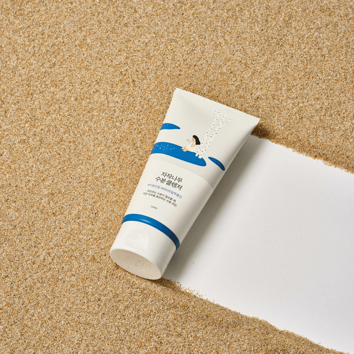 Eine Tube Birch Juice Moisturizing Cleanser liegt auf sandigem Boden mit einem weißen Streifen und zeigt auf der Verpackung eine Schwimmerillustration.