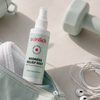 Eine Flasche Skinlick Redness Relief Mist in einem offenen Beutel, umgeben von einer Hantel und Ohrhörern.