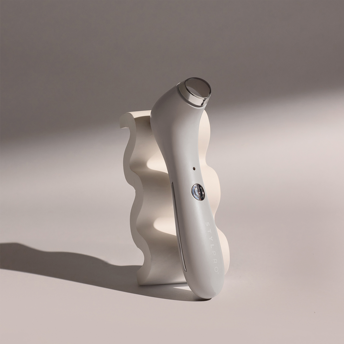 Das STYLPRO-Gesichtsmassagegerät mit Kühlfunktion in elegantem Weiß steht aufrecht auf einem wellenförmigen weißen Sockel in sanfter Beleuchtung.