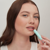 Eine Frau mit langen braunen Haaren trägt SWEED Air Blush Cream auf und blickt dabei vor einem schlichten Hintergrund in die Kamera.
