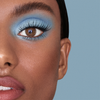 Nahaufnahme eines Gesichts mit „No Lash-Lash“ von SWEED, blauem Lidschatten, braunen Augen, rosa Lippen und langen Wimpern vor blauem Hintergrund.