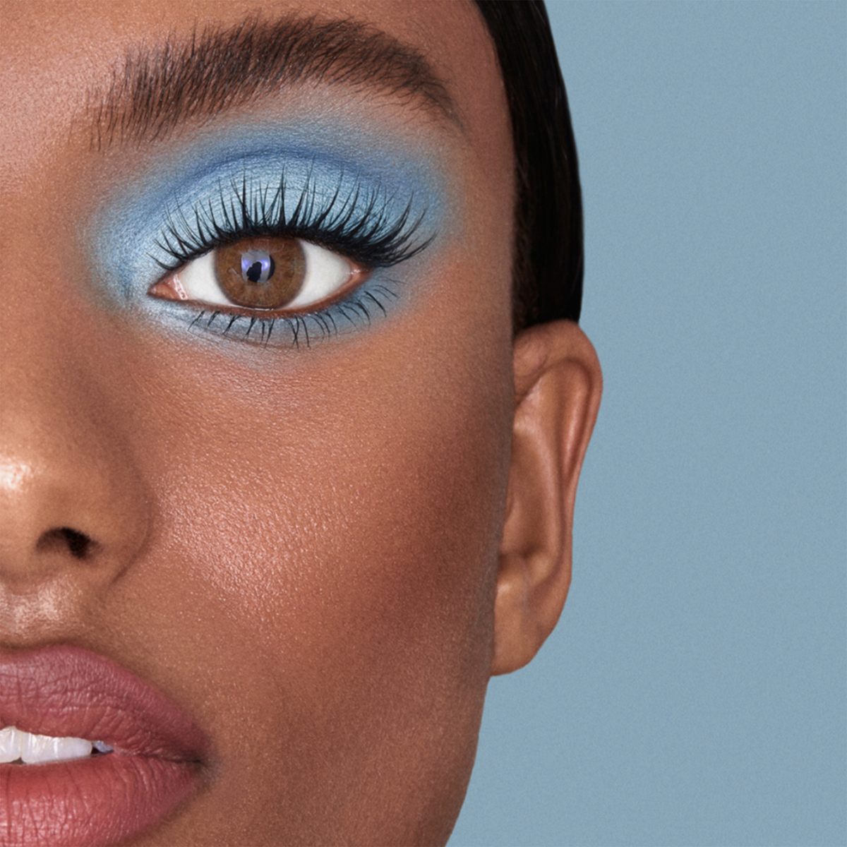 Nahaufnahme eines Gesichts mit „No Lash-Lash“ von SWEED, blauem Lidschatten, braunen Augen, rosa Lippen und langen Wimpern vor blauem Hintergrund.