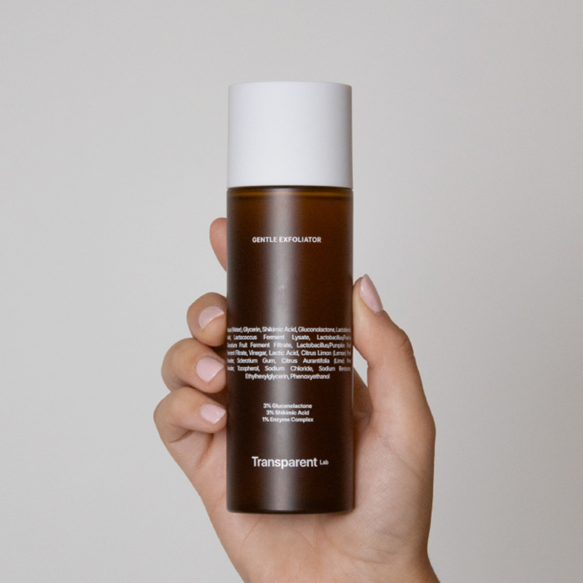 Eine Hand hält die braune Flasche des Gentle Exfoliator von Transparent Lab vor einem schlichten Hintergrund.