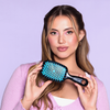 Frau mit langen braunen Haaren hält eine blau-schwarze UNbrush Detangling Hair Brush, trägt rosa, auf einem lila Hintergrund.