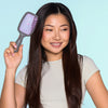 Eine lächelnde Frau mit langen dunklen Haaren hält die dunjelgraue UNbrush Detangling Hair Brush mit rosa Borsten an ihren Kopf.