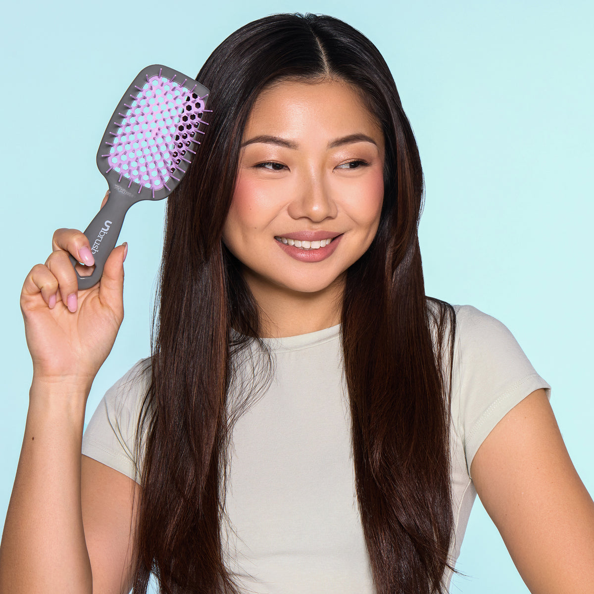 Eine lächelnde Frau mit langen dunklen Haaren hält die dunjelgraue UNbrush Detangling Hair Brush mit rosa Borsten an ihren Kopf.