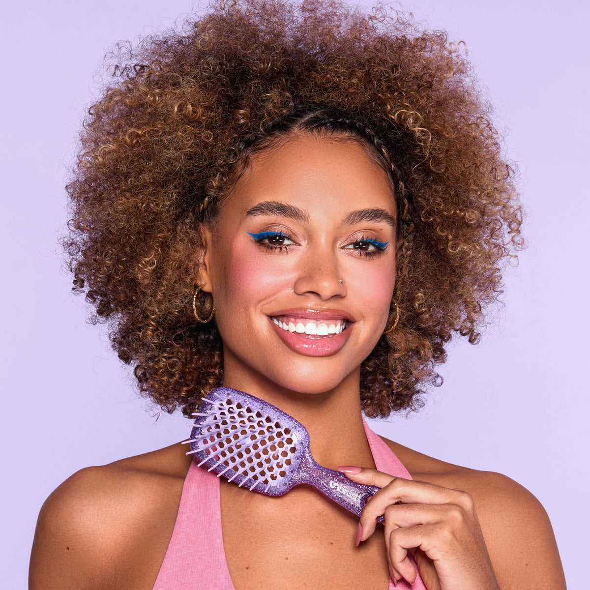 Eine lächelnde Frau mit lockigem Haar hält das eine lila Bürste aus dem UNbrush Glitter Duo Set an ihre Schulter und steht vor einem lila Hintergrund.
