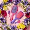 UNbrush- und Super-Mini-Set mit Haarbürsten in Neon-Pink und passenden Mini-Schlüsselanhänger-Bürsten, angeordnet in einem Kranz aus bunten Blumen.