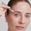 Eine Frau zupft sich mit der XLASH Cult Classic Precision Tweezer die Augenbrauen und schaut dabei direkt in die Kamera.