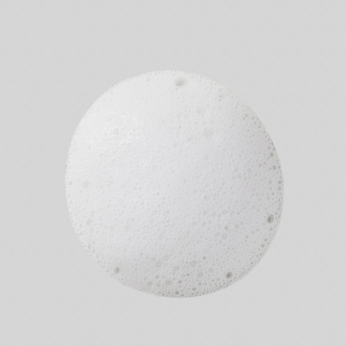 Ein runder Fleck aus weißen Schaumbläschen des Rice Enzyme Brightening Cleansing Powder liegt auf einem hellgrauen Hintergrund.