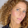 Eine Frau mit lockigem, hellbraunem Haar und blauen Augen trägt „2 IN 1 IT’S UP TO YOU LIP OR CHEEK“ von BLONDESISTER, goldene Ohrringe und ein schwarzes Oberteil.