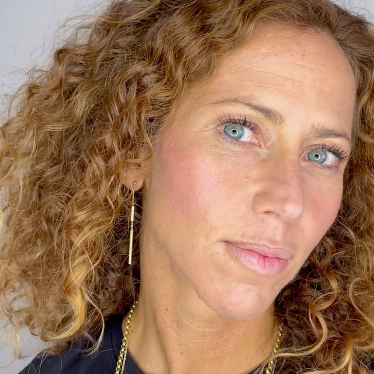 Eine Frau mit lockigem, hellbraunem Haar und blauen Augen trägt „2 IN 1 IT’S UP TO YOU LIP OR CHEEK“ von BLONDESISTER, goldene Ohrringe und ein schwarzes Oberteil.